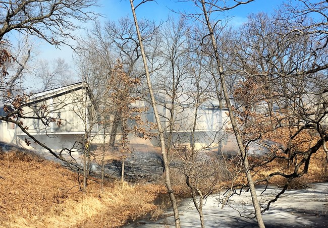 Fire adjacent to concrete building with woods and pond in foreground.