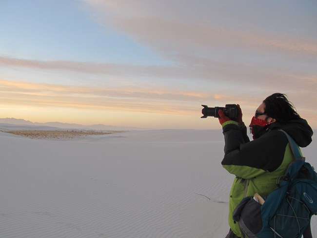 Person wearing bandana over face holds a camera at sunset.