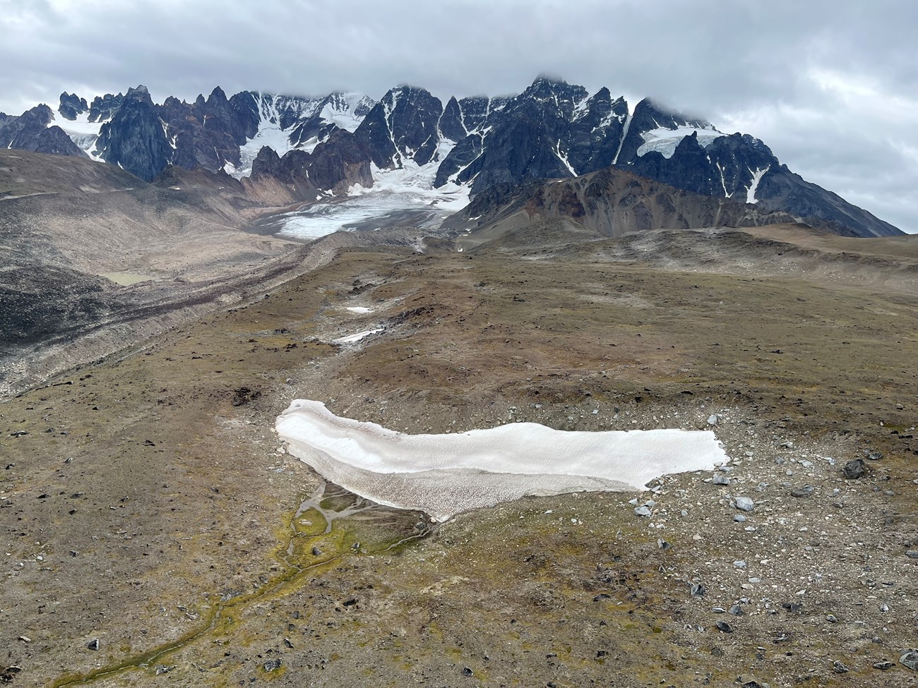 Aerial view of a long snow patch on a rocky hillside. Tall, jagged mountain peaks are in the distance