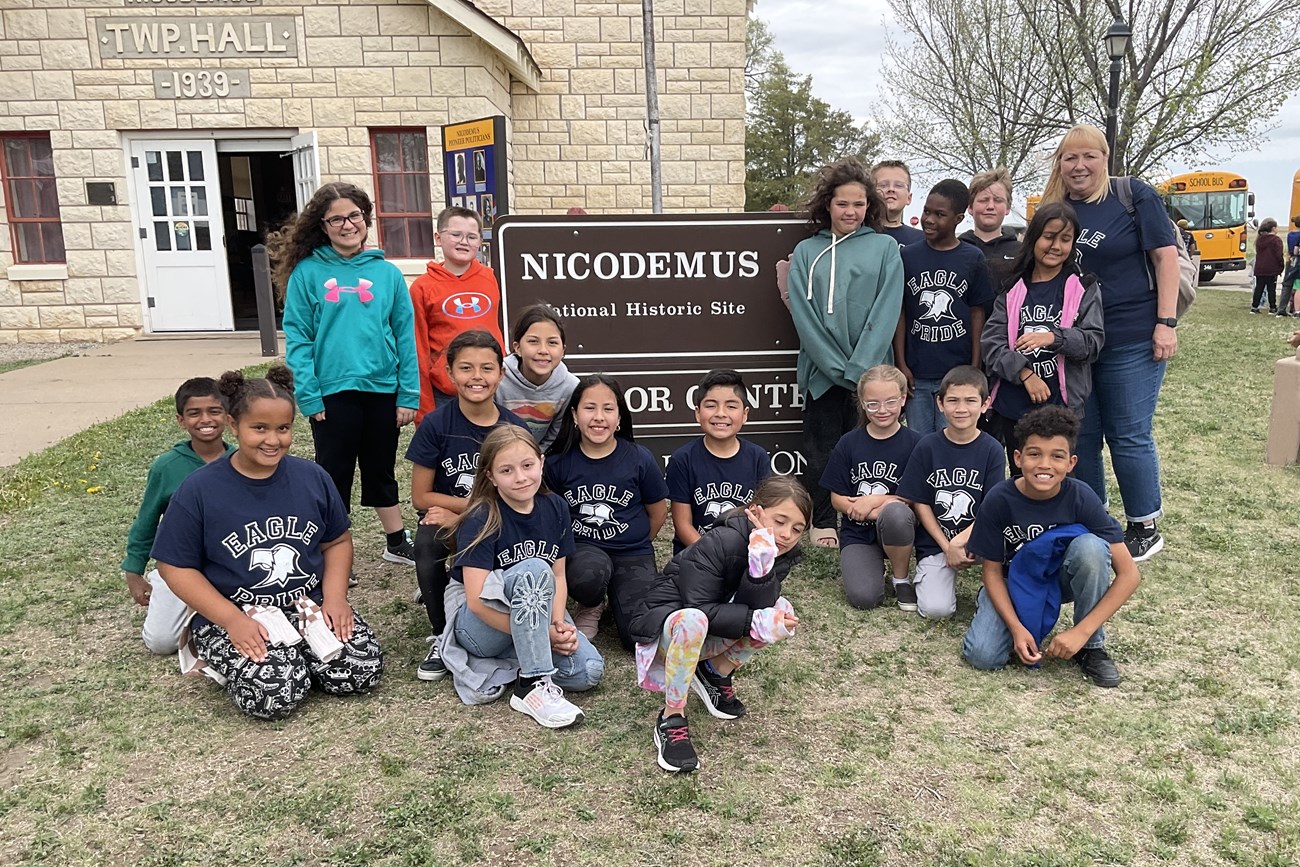 Small group of students and a teacher smiling in front of a brown sign. The children are either kneeling or standing, and most are wearing navy blue shirts with white outlined block letters that read "EAGLE PRIDE" and a white image of an eagle.