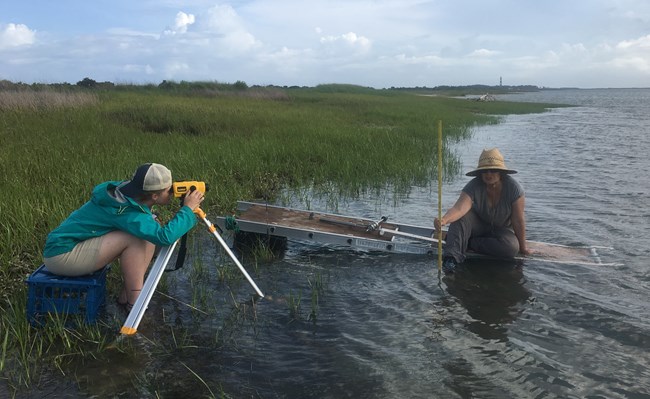 Anna Hilting holds a measuring rod sitting on a platform in the water and Elaina Sherman collects elevation data.