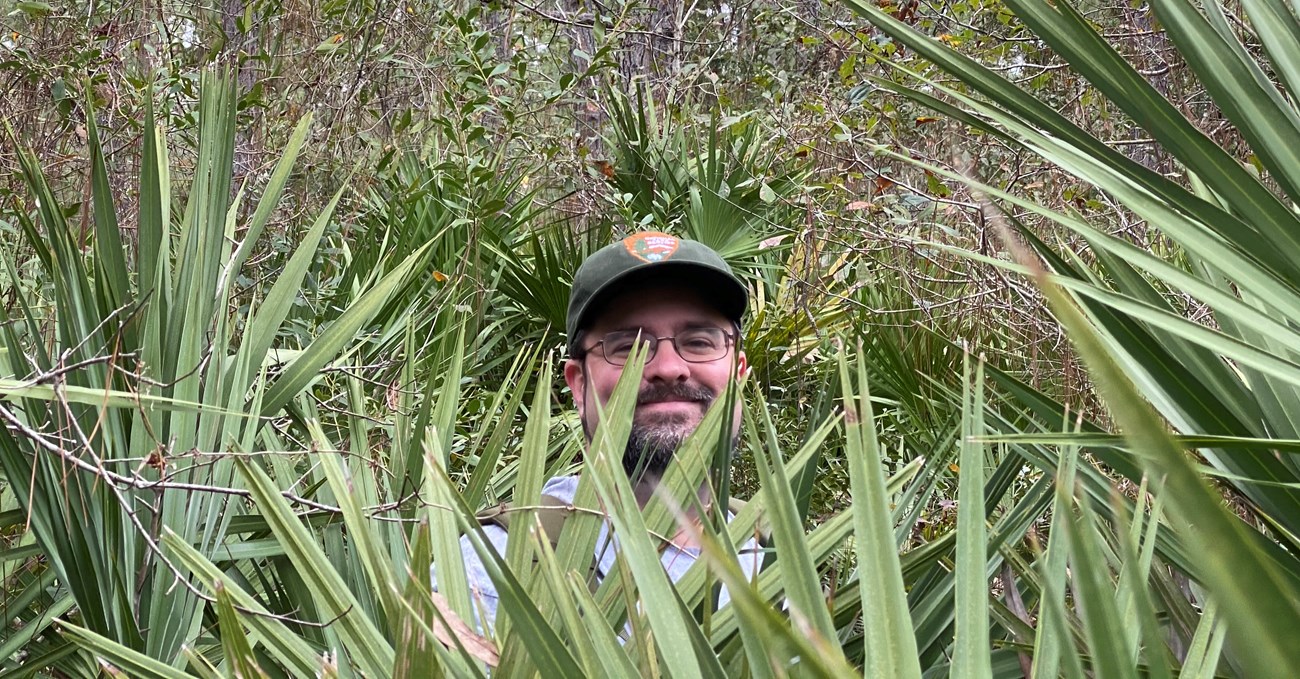 Man in hat and glasses standing in bushes. ALl you can see is his face.