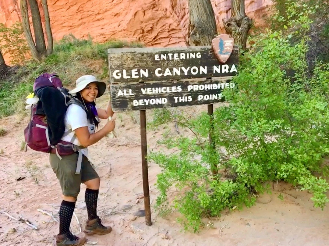 young woman dressed in hiking clothing and gear standing next to a sign that says "Entering Glen Canyon NRA"