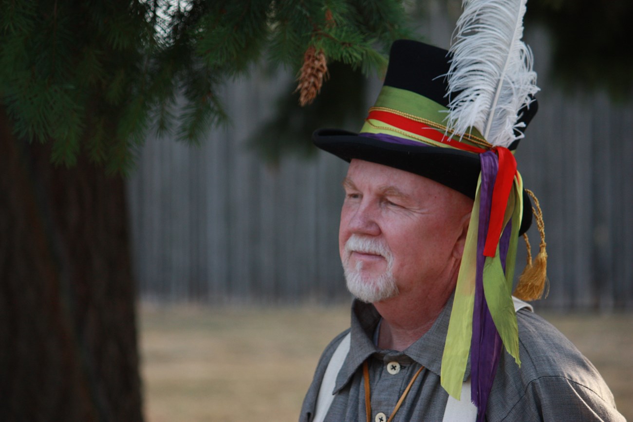 A man wears a black beaver hat decorated with colorful ribbons and a white feather.