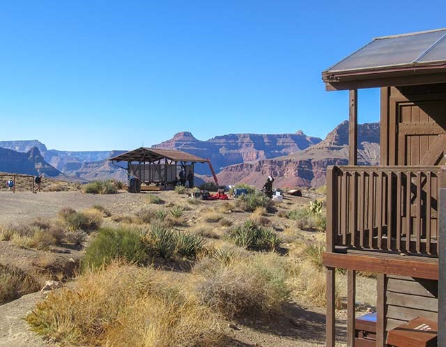 two buildings, an open-sided shade structure, and a composting toilet in a desert environment, with colorful cliffs in the distance