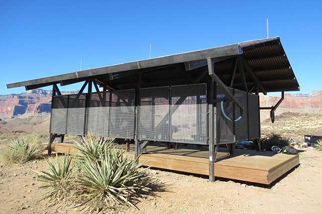 open-sided, trail-side shade structure measuring 12 x 24 feet with a corrugated metal roof and a wooden floor.