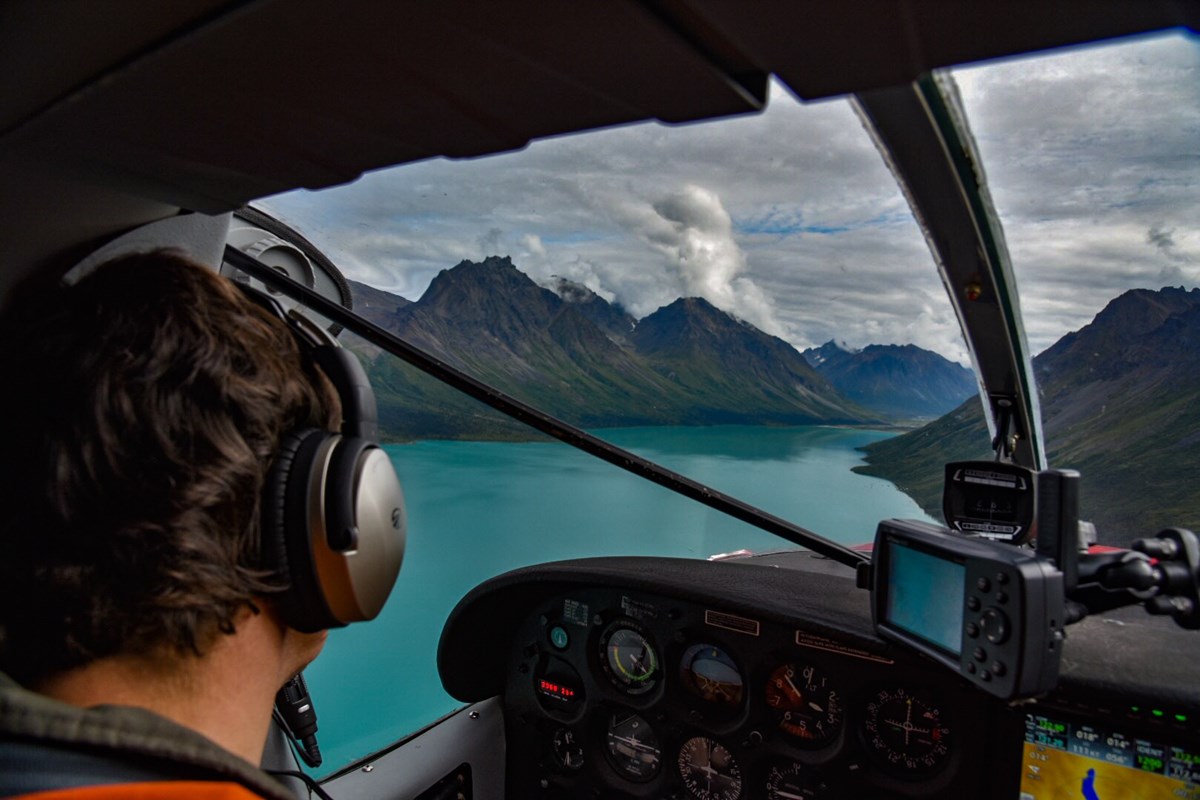 A pilot sits in a plane cockpit looking through a window to a turquoise colored lake