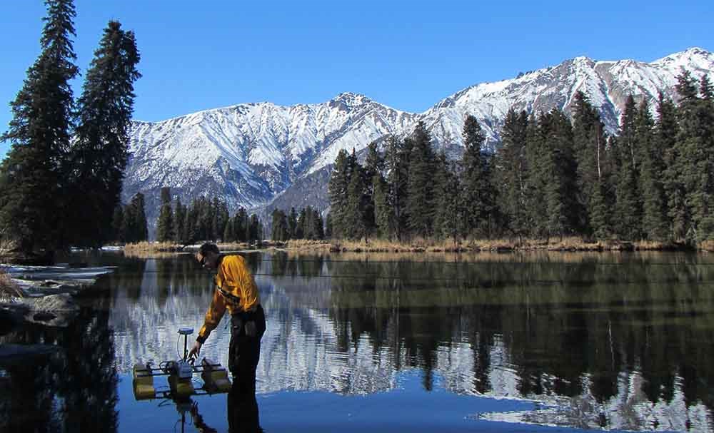 IMG_2276_web A researcher collects water discharge measurements at a lake with mountains in the background.