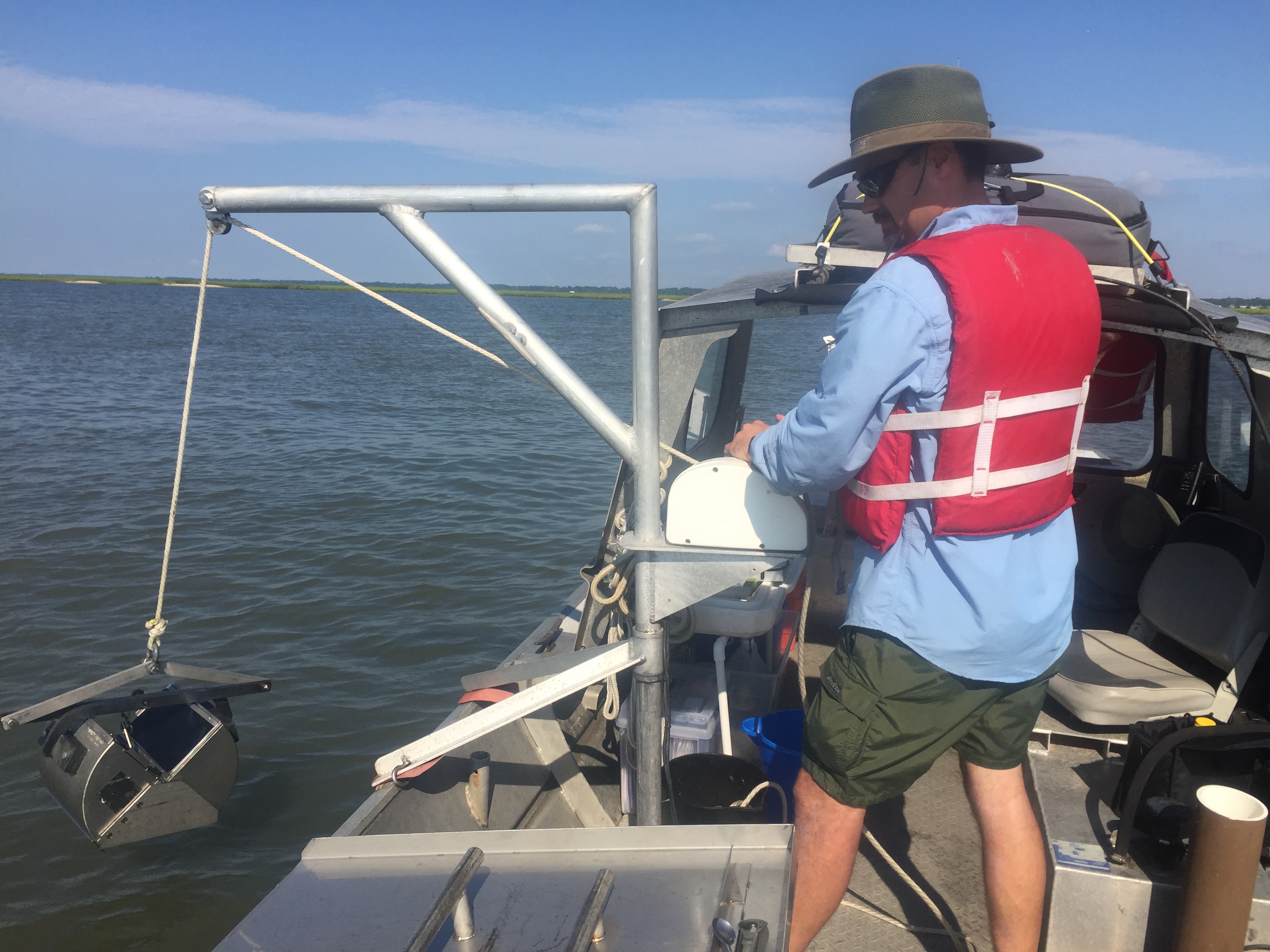 Man standing on a boat watching a boom lower a grab sampler into the water.