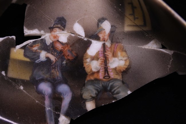 A close up image painted on porcelain, two men in funny hats playing an oboe and violin.