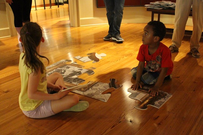 Two children sit on a floor and assemble a 4 by 8 foot puzzle