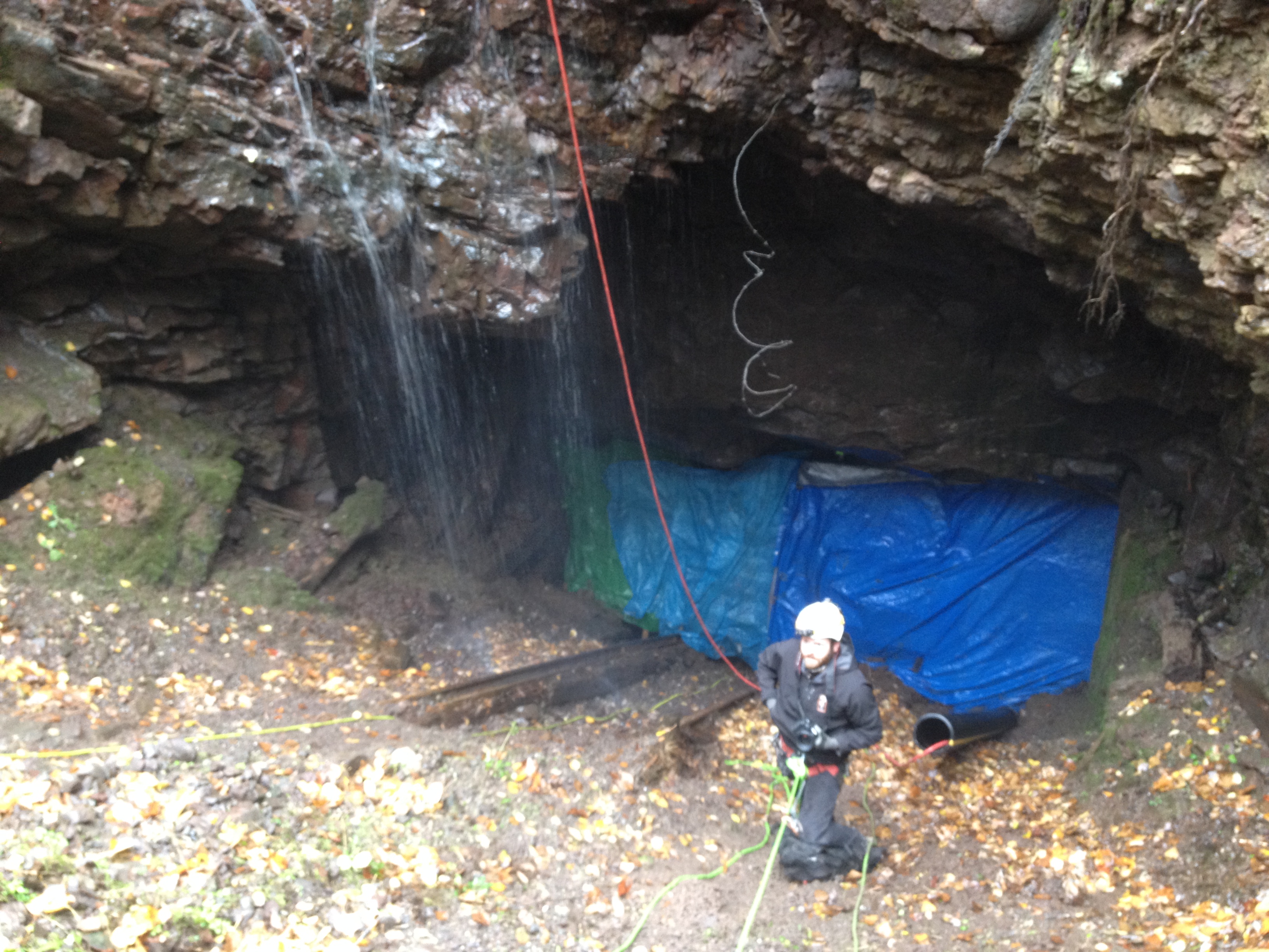 A man with safety gear underneath a rock overhang with water tripping off of it