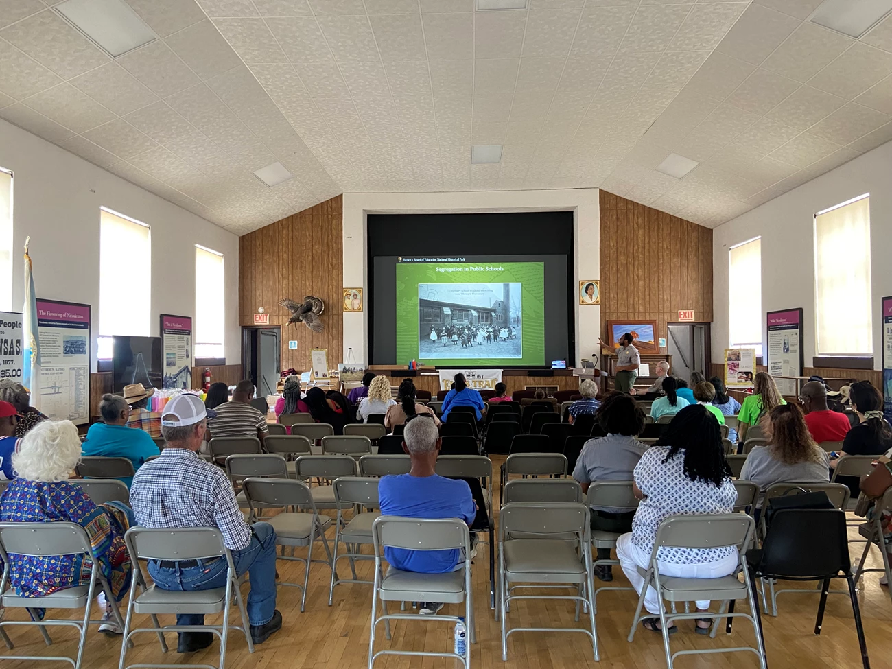 Brown v. Board of Education NHS presented at Nicodemus in 2024. Back of a crowd of people sitting in folding chairs and watching a ranger presentation on a large projector screen. The image on the projector screen is a slide from a powerpoint. The slide is green and contains the title "Segregation in Public Schools".