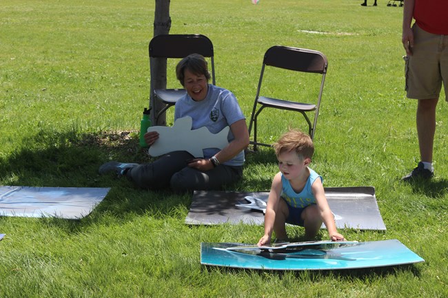 A young boy plays a game, placing a cut out of a shark over a picture of a shark while a woman sits on the ground behind him smiling