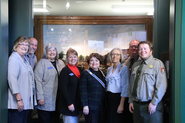 Members of the Daughters of the American Revolution and Homestead NM staff stand in front of window displaying the conserved painting on display at the park.