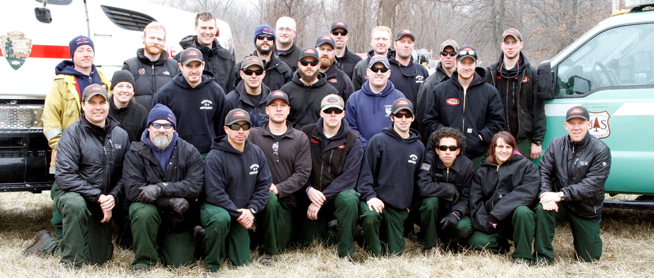 Fire staff from Indiana Dunes National Park and Midewin National Tallgrass Prairie. A group of people stand and kneel between a vehicle from the National Park Service and a vehicle from the U.S. Forest Service.