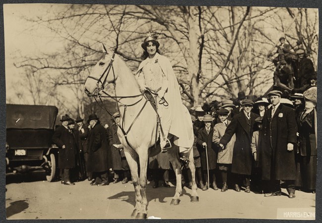 Photograph of Inez Milholland, full-length, facing forward, wearing white robes and a crown, astride a white horse in front of a crowd on a street in Washington, D.C.