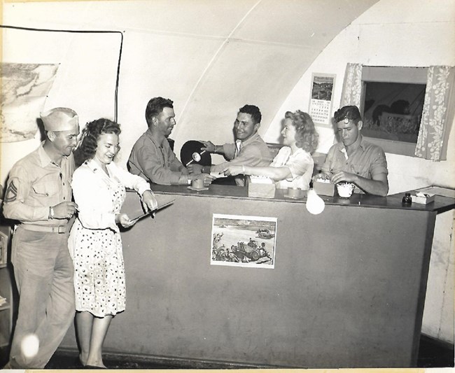 People standing behind an information desk, including three men in military uniforms, and two women in civilian clothes. One of the men is holding a record, and one of the women is pointing to something on the desk.
