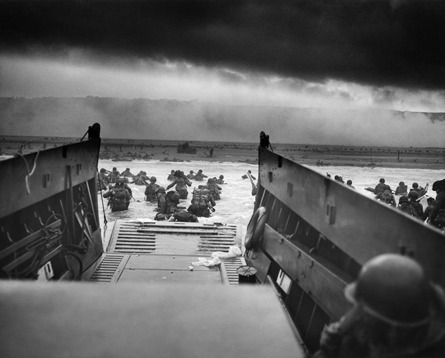 A black and white photo shows soldiers walking through water towards a beach. The photo is taken from inside a boat known as a landing craft. The metal floor and walls of the landing craft are seen in the foreground of the photo.