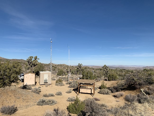 Two small buildings, a wooden stand, and two tall poles sit among scrubby desert habitat in Joshua Tree National Park. In the background are rolling hills of desert habitat with Joshua trees and blue skies with a few wispy clouds.