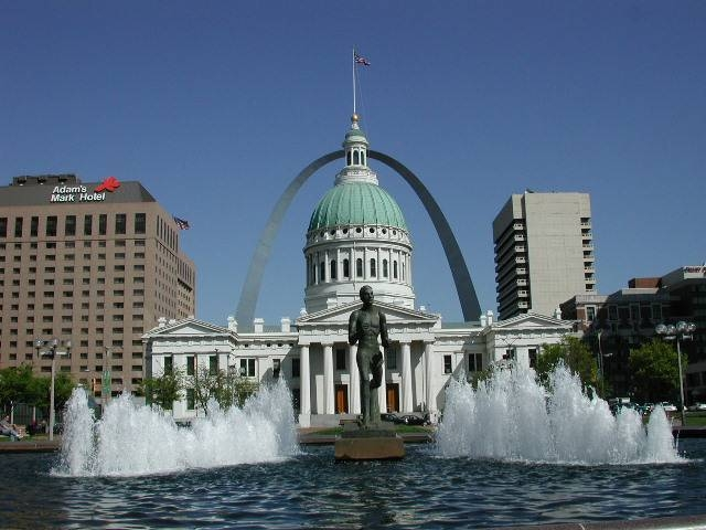 Large white, stone building with a green dome and the St. Louis Arch in the background.