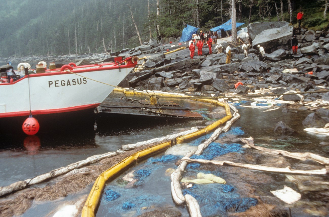 A boat next to shore where several people are involved in clean up efforts removing soil from a rocky beach