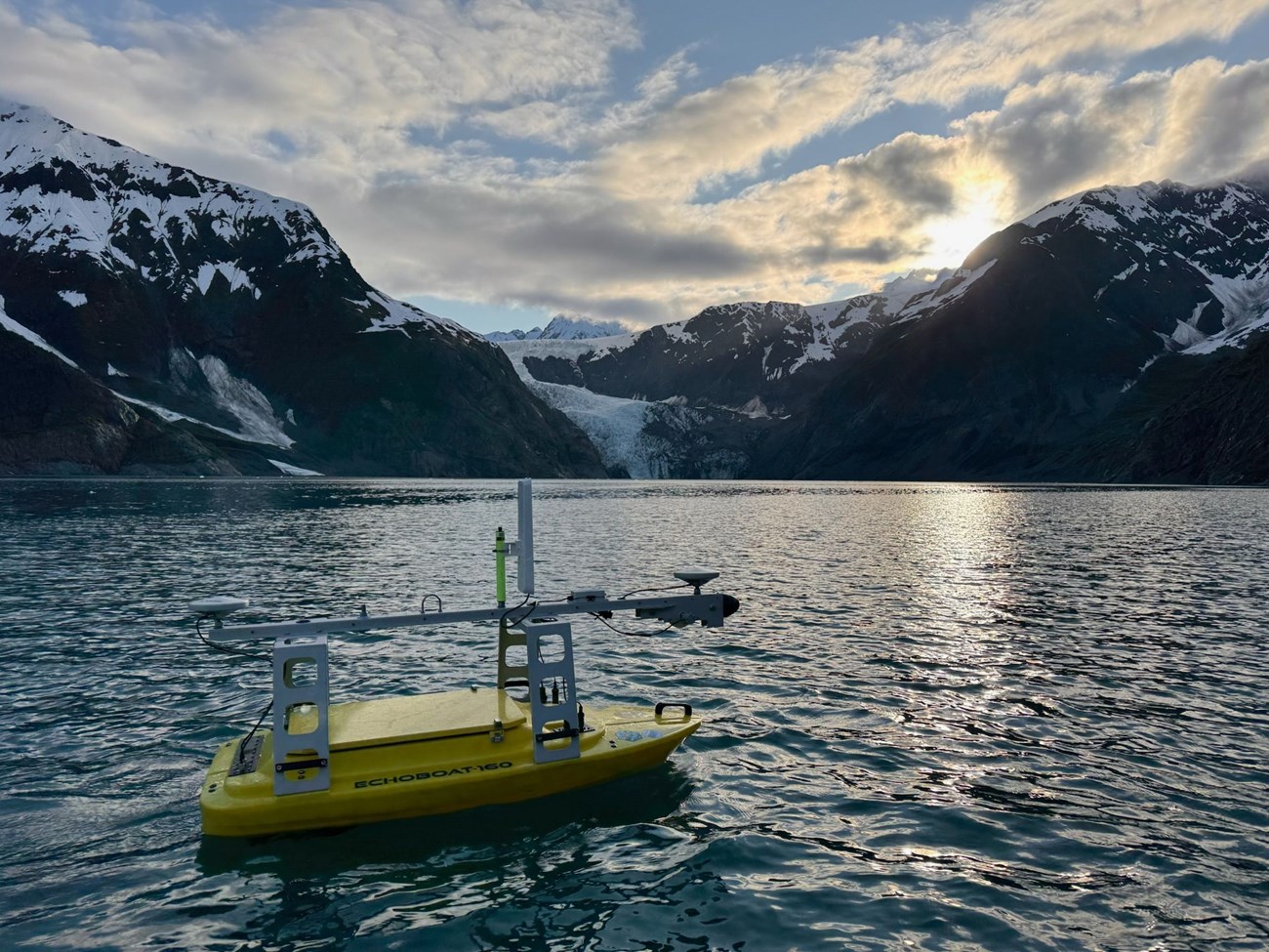 A small yellow boat floats in the water in front of a mountain range with a glacier.