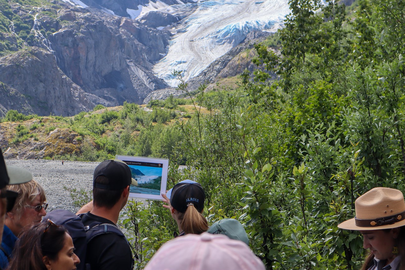Two people compare a photo of a glacier to a now smaller glacier in the distance