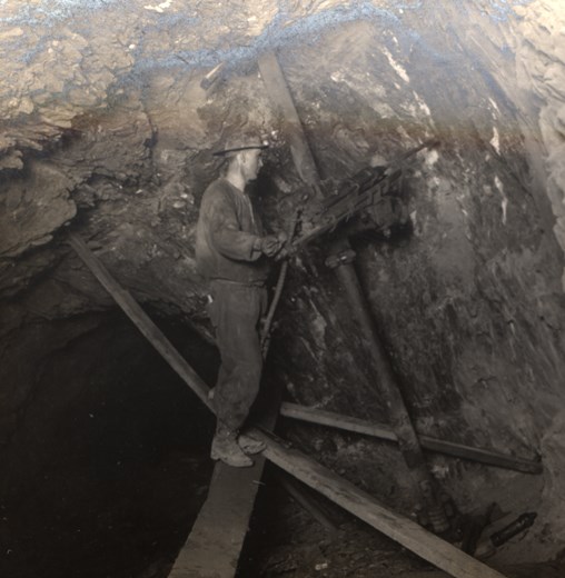 A miner stands on a wooden plank in a mineshaft.