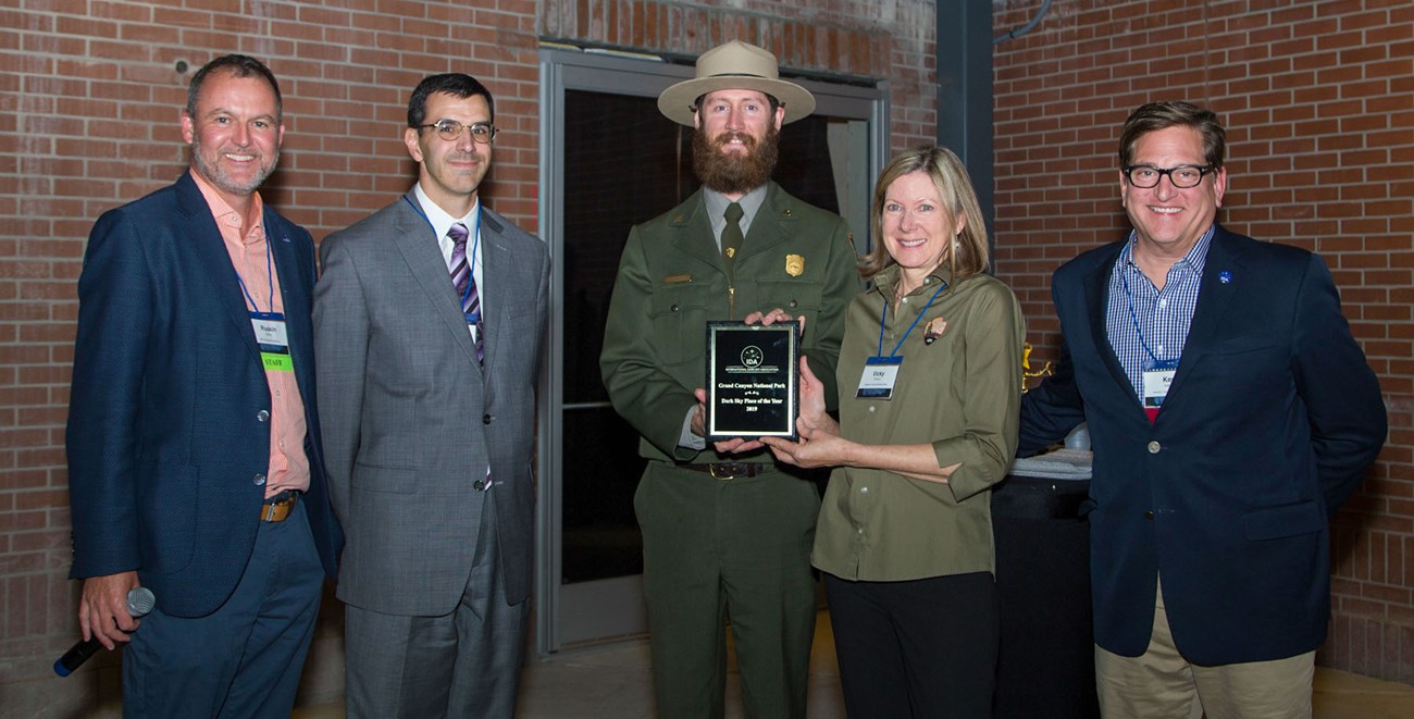 5 nicely dressed people are posing for a group photo. in the center, a park ranger and a female employee are holding an award plaque.