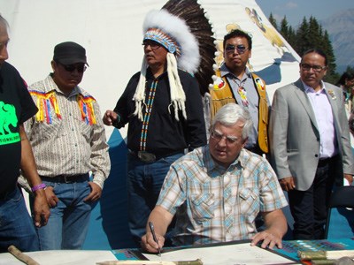 A man sits and signs the Buffalo Treaty while others in traditional indigenous attire stand behind him
