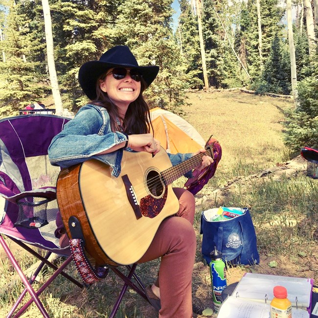 lt= Person plays an acoustic guitar while sitting on a camp chair outside.