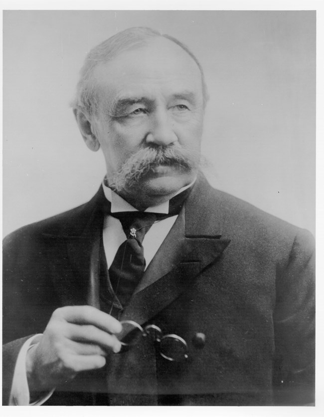 A black and white photo of a white man with a large mustache wearing a suit and holding glasses.
