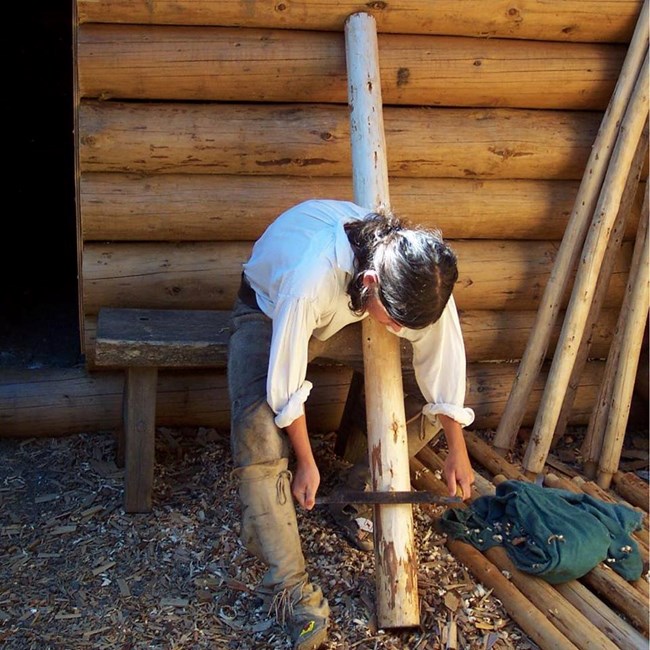 Frontal view of a ranger in costume as private and carpenter Patrick Gass seated on a bench and scraping wood off a tree pole .