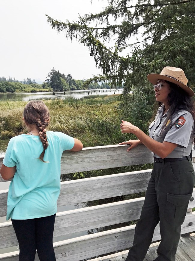 A park ranger holds her hand up as she interacts with a young girl. The two people are standing on a wooden boardwalk overlooking a wetland with many green grasses.