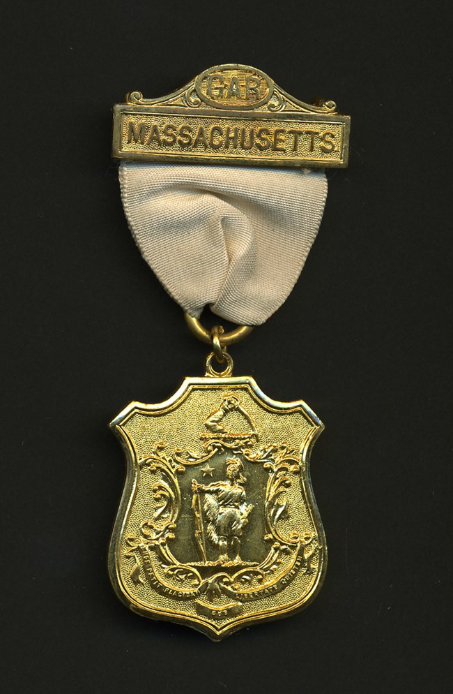 Department of Massachusetts Medal Gold-colored metal badge on off-white ribbon with seal of Massachusetts and text GAR Massachusetts on top bar