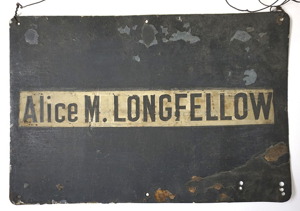 Black rectangular sign with white stripe, letters "Alice M. LONGFELLOW" at center.