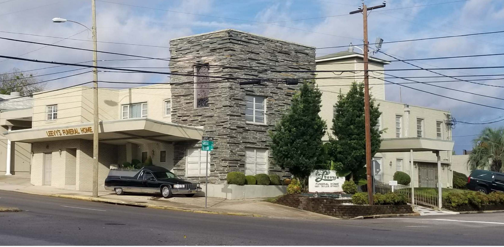 a two-story Mid-century Modern building with covered entranceways and a prominent slate-stone facade. A black hearse is parked under one of the covered entrances that displays the name of the building in gold metal lettering.