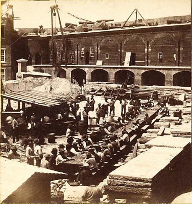Photograph of Fort Sumter's left wall being repaired. Workers seated along long table on parade ground. Temporary kitchen on parade ground with long line of men.
