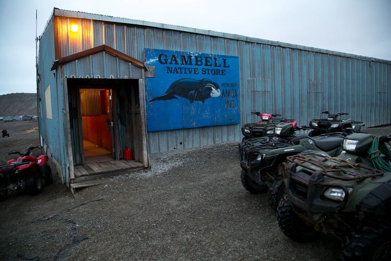 A blue rectangular building with a small entry way into a red interior is lit up with a single light. A sign read,"Gambell Native Store".