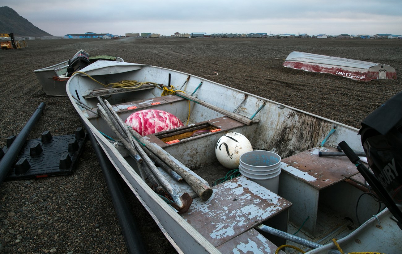 A boat lies along the beach. Inside are hunting tools, buoys, and buckets. The horizon is dotted with small houses.