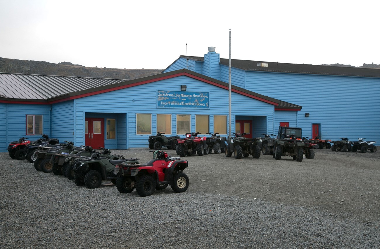 A long blue multistory building stretches across the horizon. A sign reads, "John Apangalook Memorial High School and Hugo T. Apatiki Elementary School". Numerous ATVs are neatly parked in front of the school.