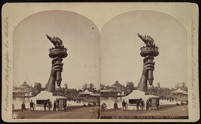 Statue of Liberty Torch A large hand holds a torch. It is mounted to a white tent in a field.