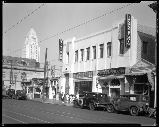 Little Tokyo A black and white photo shows a street with several storefronts. There are four 1920s automobiles parked along the street. Four girls and one man stand outside of the storefront, wearing a mix of Western and Japanese clothes.
