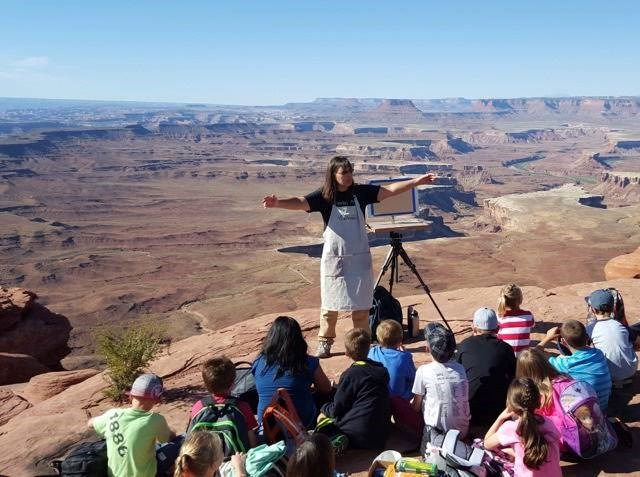 a woman stands with arms outstretched near an easel on the edge of a cliff