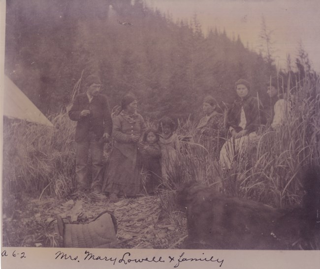 A historic photo of seven people of varying ages pose for the camera, with a forested mountain rising steeply behind them.