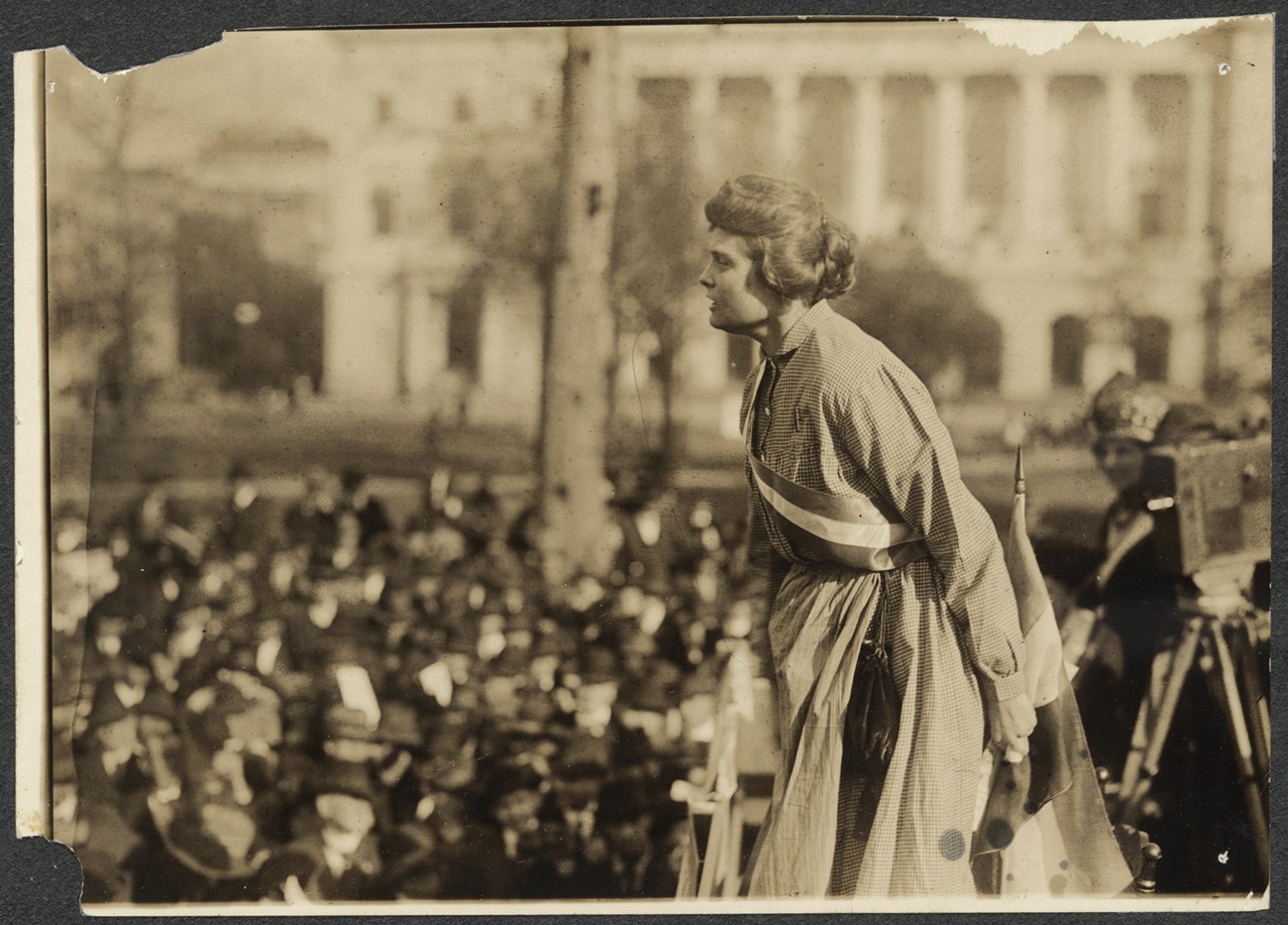 Photograph of Lucy Branham speaking at outdoor meeting during the NWP "Prison Special" tour. Branham is above a large crowd, wearing prison dress and suffrage sash, with suffrage flag and camera on tripod behind her (right).