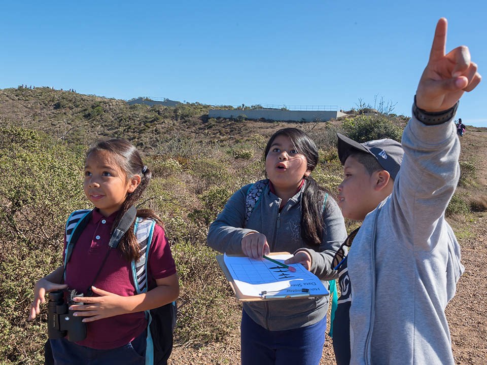 Three young students with binoculars and a data sheet pointing or looking excitedly towards the sky