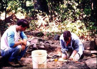 Photograph of two National Park Service archaeologists excavating the Nash Site. A woman, who is handling an artifact, and a man are both shown seated on the ground