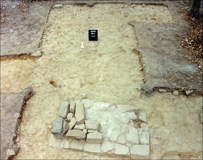 Photograph of a rectangle of chimney footing stones in a building outline. A black card near the footing marks the site.